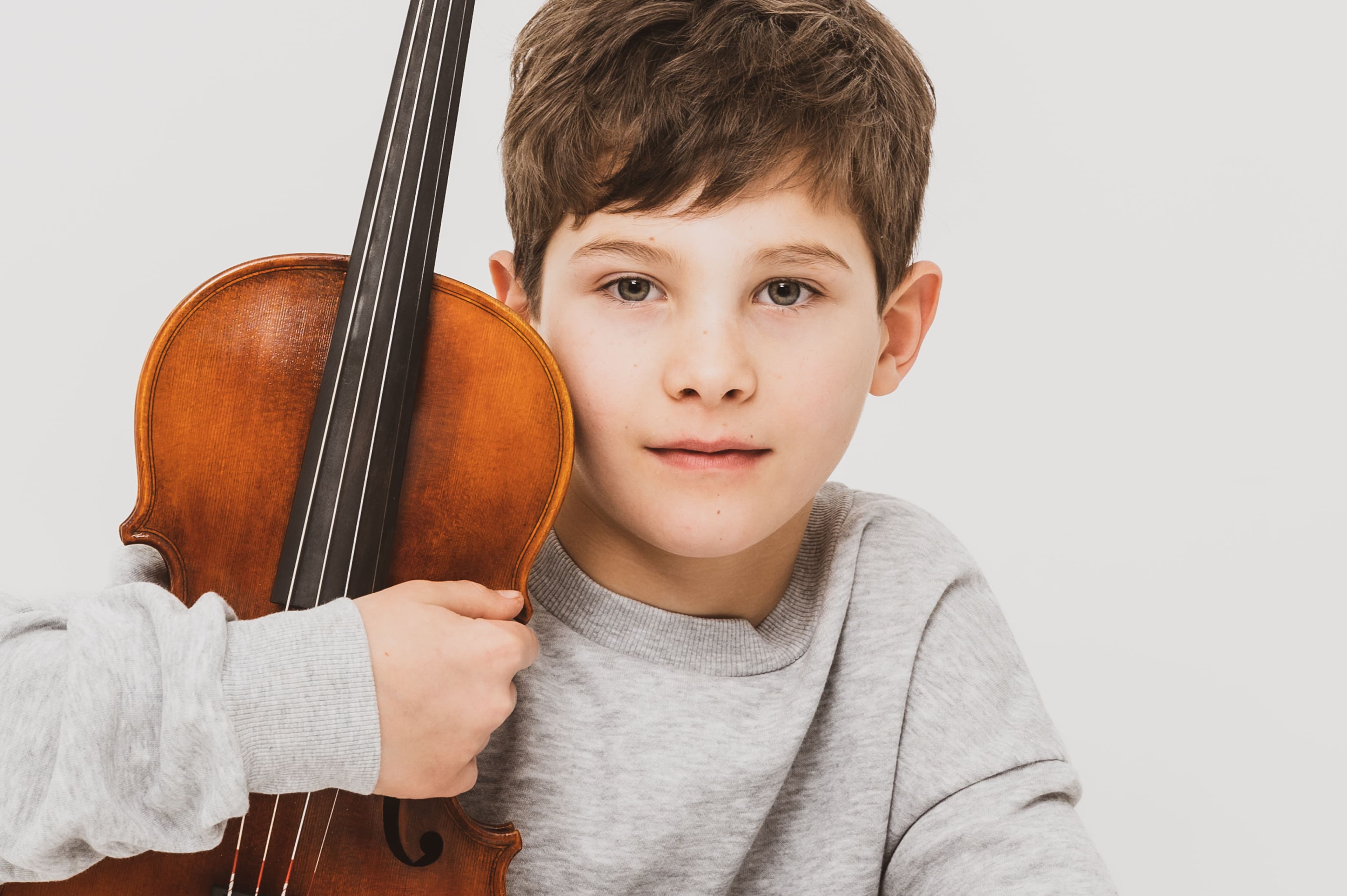 Ein Junge mit grauem Pulli sieht in die Kamera und hält im linken Arm eine Bratsche.