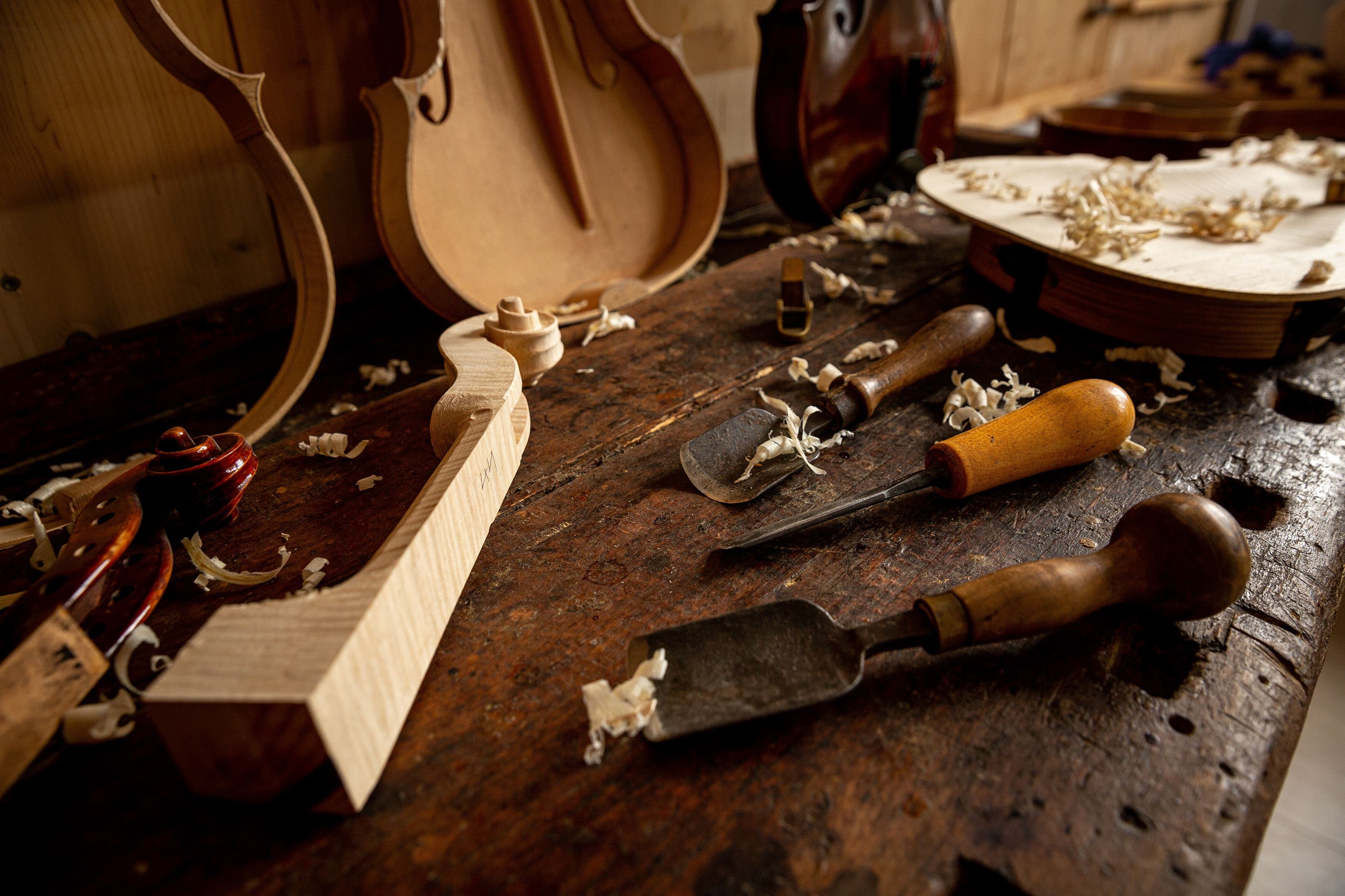 Auf einer alten Werkbank liegen verschiedene Geigenbauereisen, ein roher Geigenhals, etwas Sägespäne. Eine Geigendecke mit Bassbalken und Zargen steht an die Wand gelehnt, sowie ein vorbereiteter Zargenbogen.