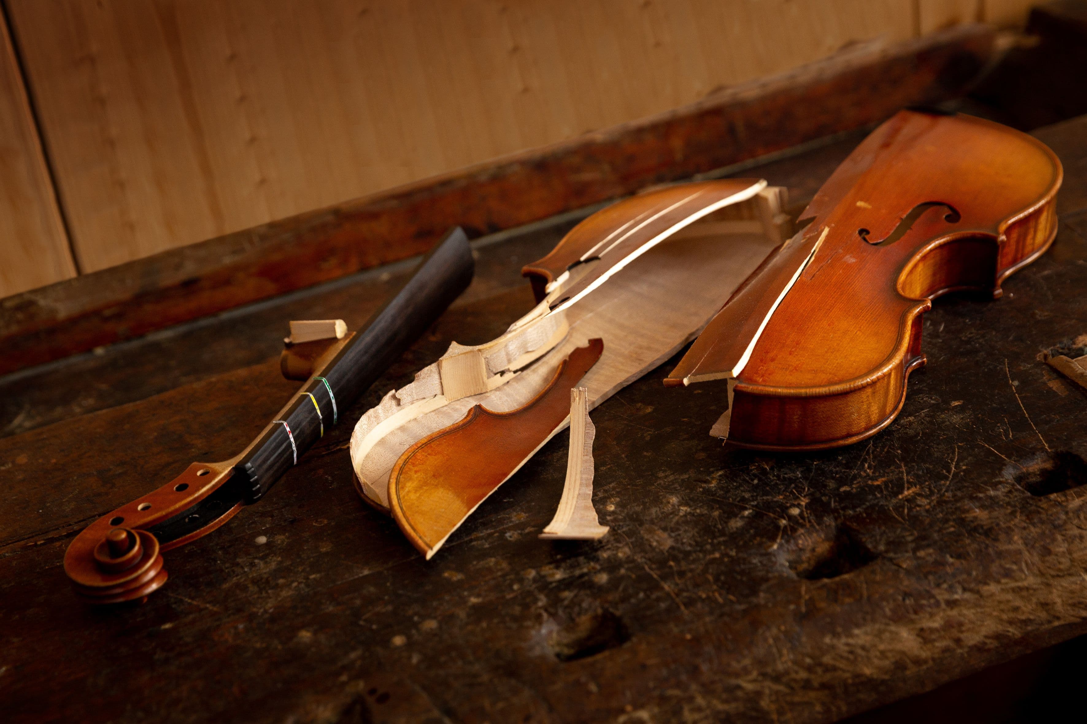 Auf einer alten Werkbank liegt eine in der Mitte durchgebrochene Geige. Der Hals der Geige liegt links daneben.