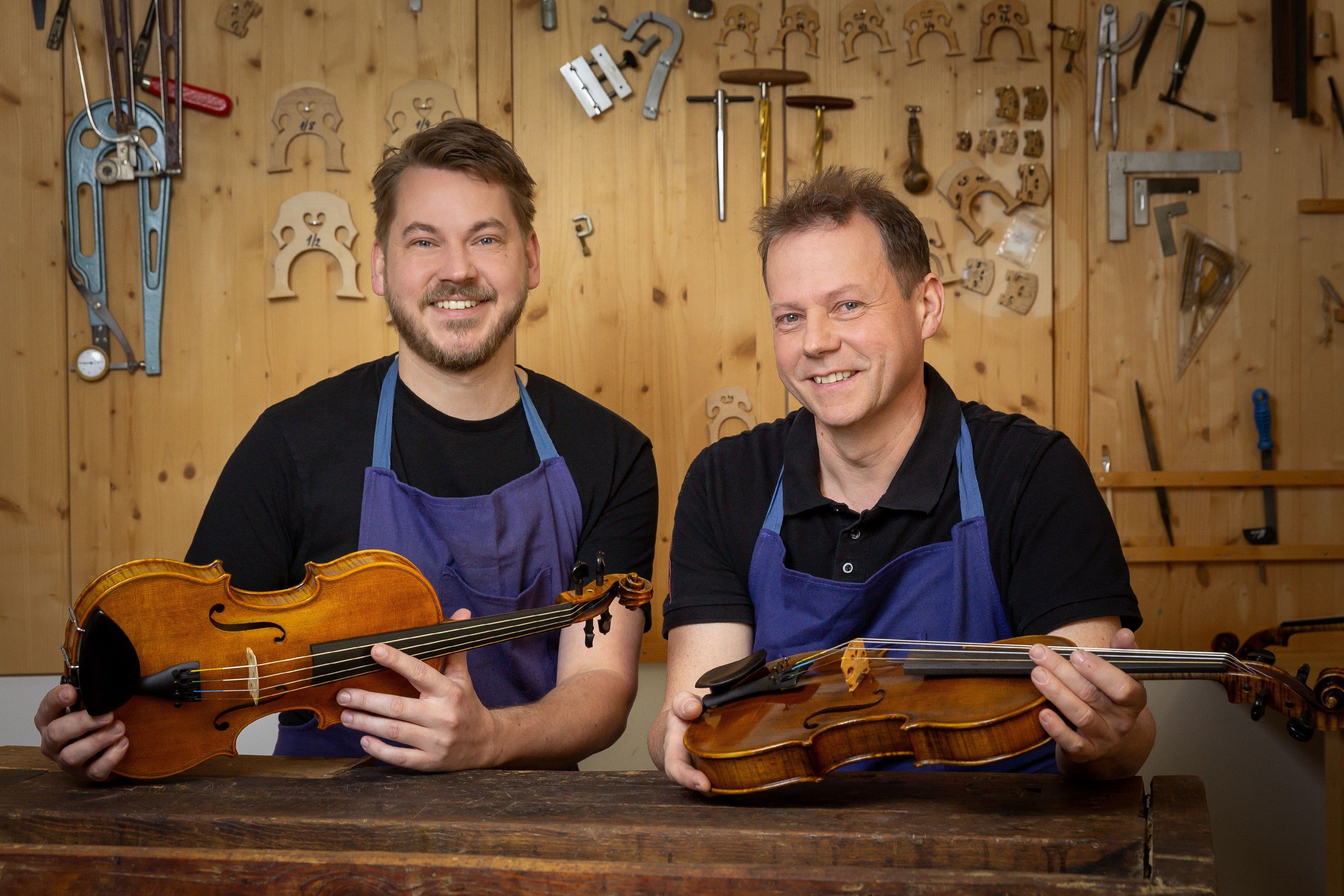 Links sitzt Michal Rácz, rechts sein Vater Petr Rácz an einer Werkbank und beide halten eines ihrer Instrumente in der Hand.