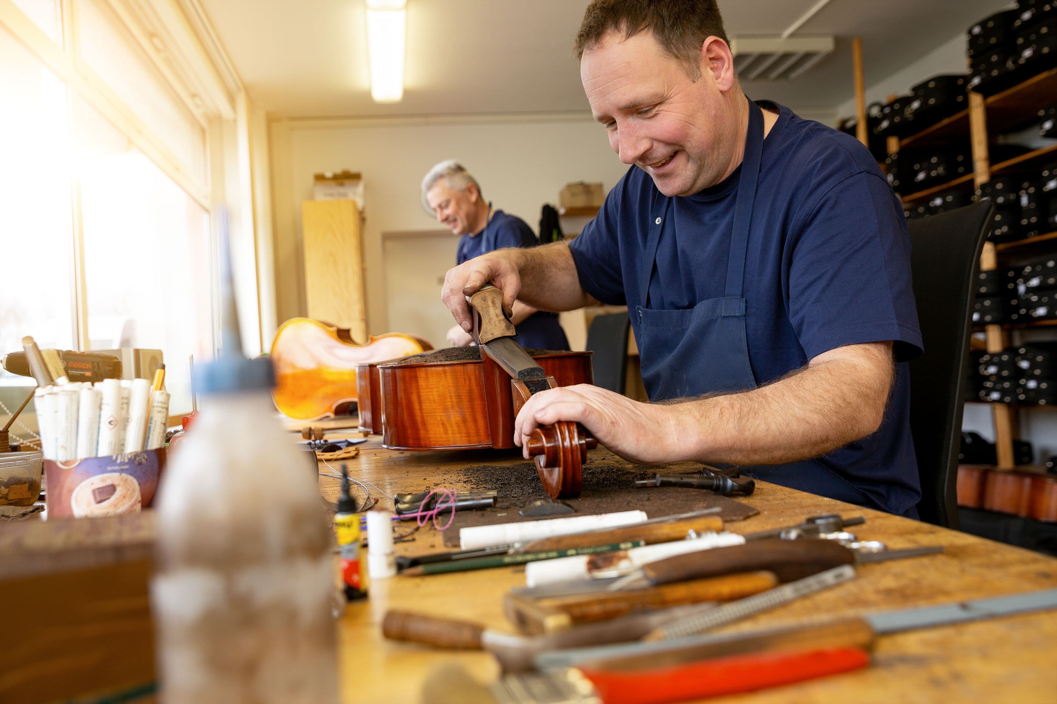 Ein Werkstattmitarbeiter sitzt gut gelaunt an seinem Arbeitsplatz, um ihn herum sein Werkzeug, und schleift ein Griffbrett ab. Im Hintergrund steht ein weiterer Mitarbeiter an einem Cello
